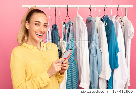 Season Of Sales And Discounts. Portrait of smiling young woman holding and using her mobile phone, chatting online or making purchases via internet standing near clothing rail at the wardrobe Season Of Sales And Discounts. Portrait of smiling young woman holding and using her mobile phone, chatting online or making purchases via internet standing near clothing rail at the wardrobe 134412900