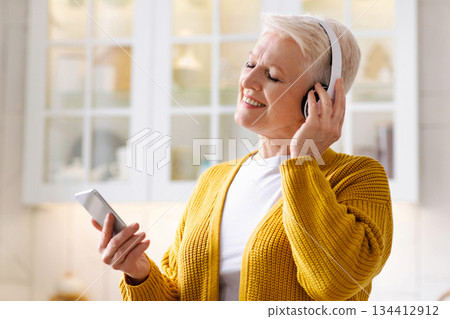 Joyful grandmother having fun at home, listening to music on mobile phone, copy space. Senior lady with closed eyes using musical mobile application on smartphone and wireless headset Joyful grandmother having fun at home, listening to music on mobile phone, copy space. Senior lady with closed eyes using musical mobile application on smartphone and wireless headset 134412912
