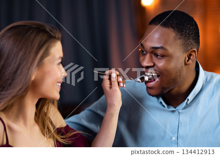 Sweet Moments. Loving Girlfriend Feeding Her Black Boyfriend With Dessert On Romantic Dinner At Home, Affectionate Interracial Couple Dining Together, Celebrating Valentine's Day , Closeup Shot Sweet Moments. Loving Girlfriend Feeding Her Black Boyfriend With Dessert On Romantic Dinner At Home, Affectionate Interracial Couple Dining Together, Celebrating Valentine's Day , Closeup Shot 134412913