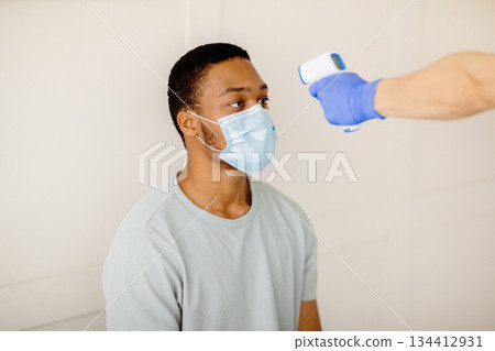 Doctor or nurse using infrared thermometer to measure temperature of black male patient before covid-19 vaccination at clinic. Medical healthcare and coronavirus immunization concept 134412931