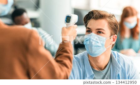 Prevention And Precautions. Woman using digital medical non-contact infrared electronic thermometer gun, measuring and screening body temperature of male student wearing surgical face mask Prevention And Precautions. Woman using digital medical non-contact infrared electronic thermometer gun, measuring and screening body temperature of male student wearing surgical face mask 134412950