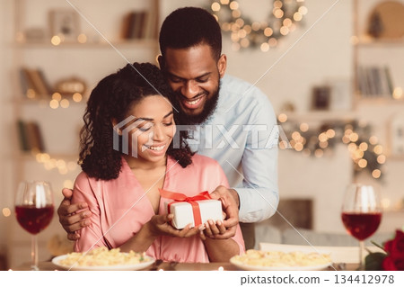 Handsome young african american guy presenting wrapped gift box to his excited girlfriend at cafe, making her special surprise. Loving couple celebrating Valentine's Day, birthday or anniversary 134412978