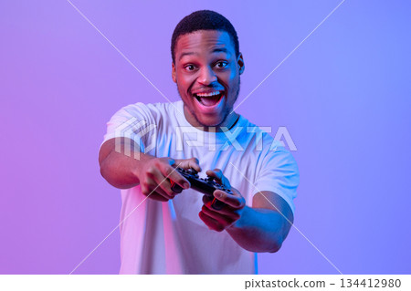 Overjoyed Black Gamer Man With Joystick Playing Video Games In Neon Lighting, Emotional Excited Guy Having Fun While Standing In Neon Lighting Over Purple Studio Background, Looking At Camera 134412980