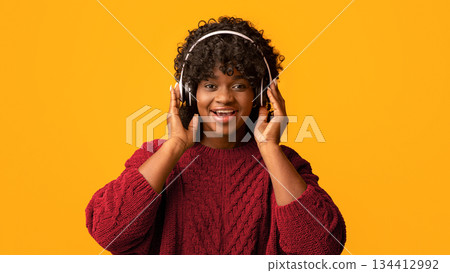 Pretty young black woman in wireless headset listening to music and smiling, african american millennial lady enjoying good sound with modern headphones, yellow studio background, panorama, copy space 134412992