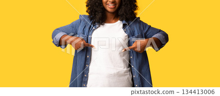 T-shirt for your print. Attractive happy african american lady in casual outfit smiling and pointing at her white blank t-shirt, posing on yellow studio background, copy space 134413006