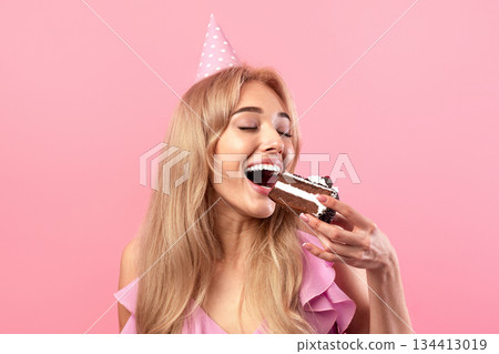 Pretty young woman eating yummy cake, wearing birthday cap, celebrating holiday on pink studio background. Sexy millennial blonde enjoying delicious festive dessert on B-day party Pretty young woman eating yummy cake, wearing birthday cap, celebrating holiday on pink studio background. Sexy millennial blonde enjoying delicious festive dessert on B-day party 134413019