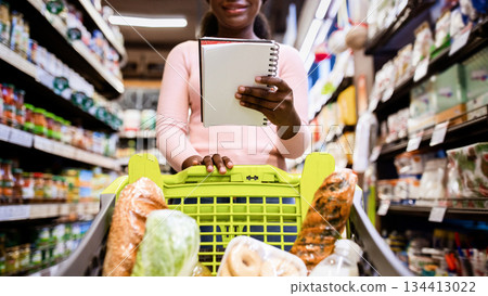 Young black woman with shopping cart full of products looking into her grocery list at supermarket. Lovely African American lady buying products at mall, low angle view. Selective focus Young black woman with shopping cart full of products looking into her grocery list at supermarket. Lovely African American lady buying products at mall, low angle view. Selective focus 134413022