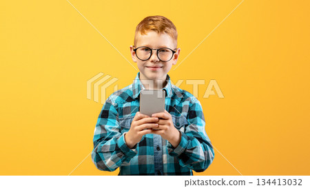 Smiling boy schooler playing game or using amazing application on smartphone over yellow studio background, copy space. Happy ginger kid in glasses using newest mobile phone, entertainment concept Smiling boy schooler playing game or using amazing application on smartphone over yellow studio background, copy space. Happy ginger kid in glasses using newest mobile phone, entertainment concept 134413032