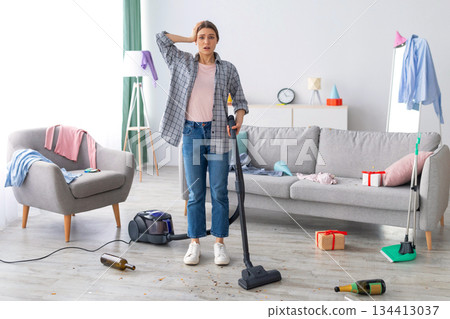 After party chaos. Dismayed millennial woman standing in her messy apartment with vacuum cleaner, holding head in terror, copy space. Terrified female student cleaning dirty university dorm room 134413037
