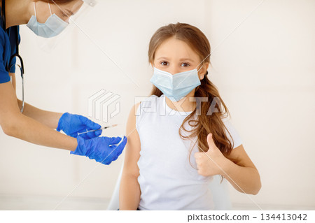 Little girl in face mask gesturing thumb up while receiving covid-19 vaccine injection at health centre. Cute child approving of coronavirus immunization, being protected against global virus Little girl in face mask gesturing thumb up while receiving covid-19 vaccine injection at health centre. Cute child approving of coronavirus immunization, being protected against global virus 134413042