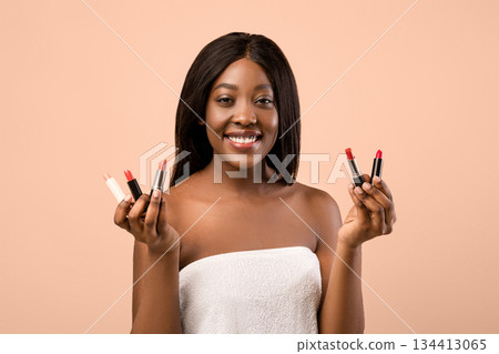 Closeup of smiling beautiful young black woman wrapped in towel holding two lipsticks in her hands, posing over beige background. Professional cosmetics makeup, lipstick, lip-gloss concept Closeup of smiling beautiful young black woman wrapped in towel holding two lipsticks in her hands, posing over beige background. Professional cosmetics makeup, lipstick, lip-gloss concept 134413065