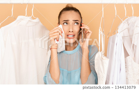 I Have Nothing To Wear. Portrait of upset stressed woman talking on mobile phone and crying, wiping tears with shirt, having panic, standing in rack of clothes between hangers I Have Nothing To Wear. Portrait of upset stressed woman talking on mobile phone and crying, wiping tears with shirt, having panic, standing in rack of clothes between hangers 134413079