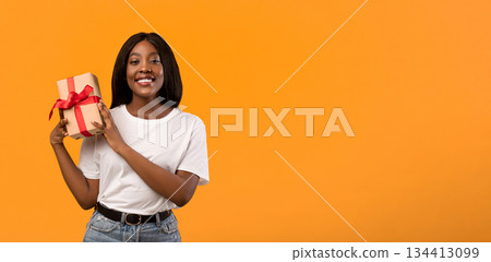 Happy african american young woman holding gift box next to her ear, wondering what is inside, orange studio background, panorama with copy space. Birthday gift, present, surprise concept 134413099