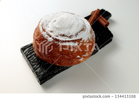 Round, delicious bun with cinnamon and powdered sugar on a black wooden board on a white background Round, delicious bun with cinnamon and powdered sugar on a black wooden board on a white background 134413108