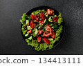 Vegetable salad of tomatoes, cucumbers, onions, lettuce dressed with aromatic dressing in a black plate on a gray background 134413111