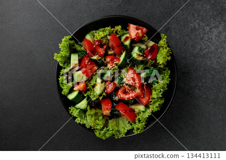 Vegetable salad of tomatoes, cucumbers, onions, lettuce dressed with aromatic dressing in a black plate on a gray background 134413111