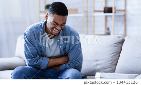 Food Poisoning. Black Man Suffering From Stomachache After Eating Pizza Touching Aching Abdomen Sitting On Couch At Home. Health Problem, Appendicitis, Abdominal Inflammation And Gastritis Concept. Food Poisoning. Black Man Suffering From Stomachache After Eating Pizza Touching Aching Abdomen Sitting On Couch At Home. Health Problem, Appendicitis, Abdominal Inflammation And Gastritis Concept. 134413126