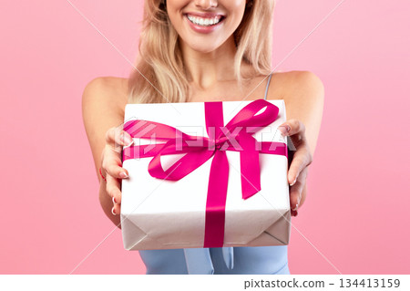 Lovely young woman giving wrapped gift box at camera over pink studio background. Cheerful millennial lady offering holiday present with smile, celebrating birthday or anniversary 134413159