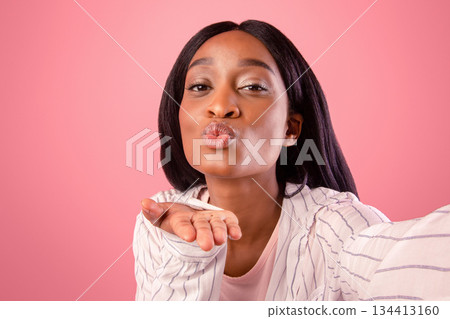 Attractive young black woman taking selfie, blowing air kiss on pink studio background. Lovely African American lady posing to mobile camera and making photo of herself 134413160
