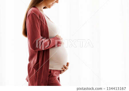 Profile Portrait Of Smiling Pregnant Woman Tenderly Embracing Belly Against Window At Home, Happy Expectant Mother Enjoying Her Healthy Pregnancy And Awaiting Time, Side View With Copy Space 134413167