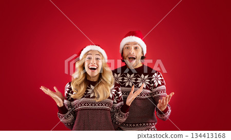 Excited millennial couple in Santa hats and Christmas sweaters on red background 134413168