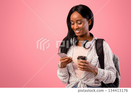 Young black lady using cellphone and holding takeout coffee on pink studio background. African American student with backpack and headphones browsing web on mobile device Young black lady using cellphone and holding takeout coffee on pink studio background. African American student with backpack and headphones browsing web on mobile device 134413176