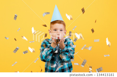 Birthday boy in paper hat blowing at sparkling confetti, having great party over yellow studio background. Happy ginger kid celebrating birthday, throwing bright confetti all over 134413181