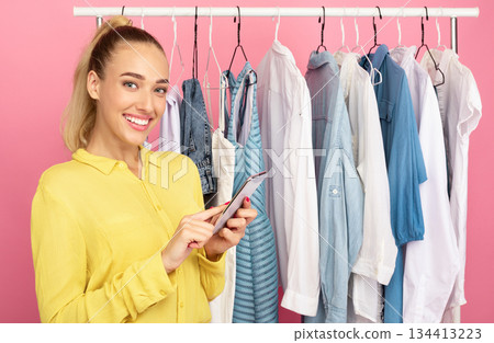 Fashion And Style. Portrait of smiling woman stylist holding and using smartphone, texting sms or making purchases in online store standing near rack with clothes over pastel pink studio background Fashion And Style. Portrait of smiling woman stylist holding and using smartphone, texting sms or making purchases in online store standing near rack with clothes over pastel pink studio background 134413223