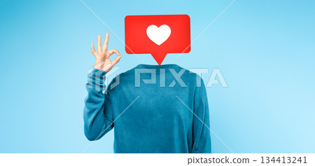 A person dressed in a blue sweater makes an OK hand gesture, with a heart icon superimposed above where their head would be. The simple blue backdrop highlights the playful social media theme. 134413241