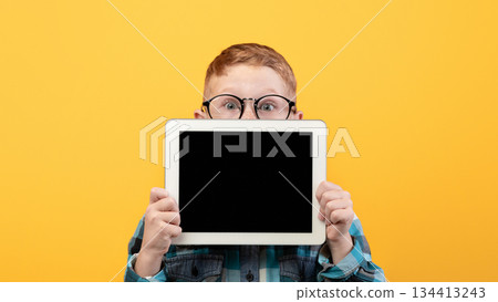 Amazed kid in eyeglasses hiding behind digital tablet with empty screen over yellow studio background. Excited redhead schoolboy showing pad, mockup, advertisement for cute application 134413243