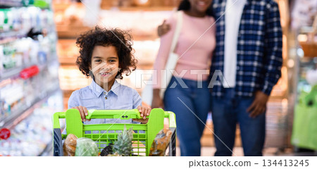 Happy little black girl with shopping trolley and her parents buying goods at modern supermarket, free space. Millennial African American family purchasing groceries at big mall Happy little black girl with shopping trolley and her parents buying goods at modern supermarket, free space. Millennial African American family purchasing groceries at big mall 134413245