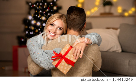 Delighted wife holding xmas gift and hugging her husband, celebrating Christmas together at home, empty space. Young woman happy to get Christmas present from her boyfriend, embracing him and smiling 134413249