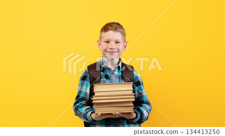 Smiling schoolboy with backpack holding heap of books, yellow studio background, copy space. Happy boy going to school, holding books, enjoying his school days. Kids education concept 134413250
