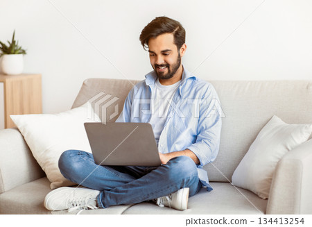 Distant Work. Happy Arab Freelancer Man Using Laptop Computer At Home, Typing On Keyboard, Young Eastern Guy Sitting On Sofa In Living Room, Working Remotely On Online Project, Copy Space 134413254
