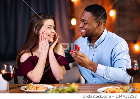 Happy African American Man Making Proposal To His Surprised White Girlfriend, Asking To Marry Him During Romantic Date Dinner In Restaurant, Giving Box With Diamond Ring, Free Space 134413258
