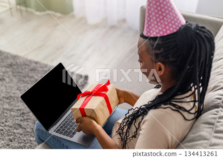 Top view of black woman in party hat using laptop with empty screen, showing gift box, copy space. African american lady having birthday party online, celebrating with friends or family Top view of black woman in party hat using laptop with empty screen, showing gift box, copy space. African american lady having birthday party online, celebrating with friends or family 134413261