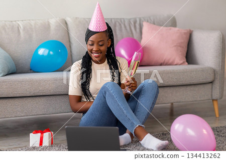 Positive african american woman in party hat using laptop at home, holding glass of champagne and smiling at computer screen, sitting in room decorated with colorful balloons and presents, copy space Positive african american woman in party hat using laptop at home, holding glass of champagne and smiling at computer screen, sitting in room decorated with colorful balloons and presents, copy space 134413262
