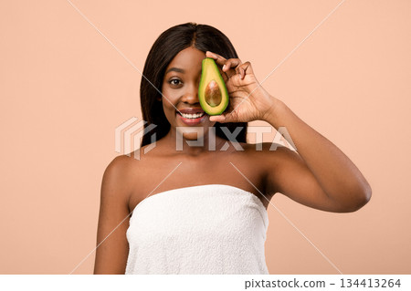 Smiling beautiful black woman with long hair wrapped in towel covering eyes with half of avocado over beige studio background. Youth, beauty, anti-aging recipes, healthy diet concept Smiling beautiful black woman with long hair wrapped in towel covering eyes with half of avocado over beige studio background. Youth, beauty, anti-aging recipes, healthy diet concept 134413264