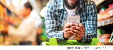 Black Man Using Mobile Coupon App For Groceries Shopping Buying Food In Supermarket, Standing With Cart In Hypermarket. African Guy Using Smartphone Purchasing Grocery In Shop. Selective Focus Black Man Using Mobile Coupon App For Groceries Shopping Buying Food In Supermarket, Standing With Cart In Hypermarket. African Guy Using Smartphone Purchasing Grocery In Shop. Selective Focus 134413402