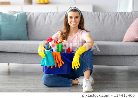 Full length portrait of happy young maid with bucket of cleaning supplies sitting on floor at living room. Cheerful millennial housewife with sanitary products smiling at camera Full length portrait of happy young maid with bucket of cleaning supplies sitting on floor at living room. Cheerful millennial housewife with sanitary products smiling at camera 134413406