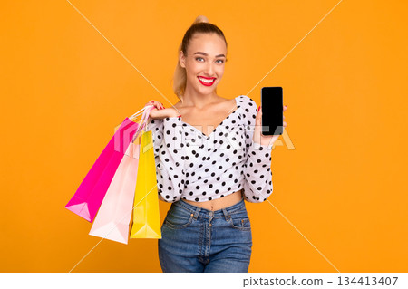 Ordering Online. Portrait of smiling woman holding colorful shopping bags and showing cell phone with blank black screen to camera, standing isolated over pastel orange studio wall background 134413407