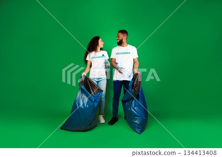 Two Diverse Volunteers With Big Blue Garbage Bags Picking Wasted Plastic Bottles Posing On Green Background. Studio Shot. Keep Environment Clean, Ecology And Volunteering Work Concept. Full Length 134413408