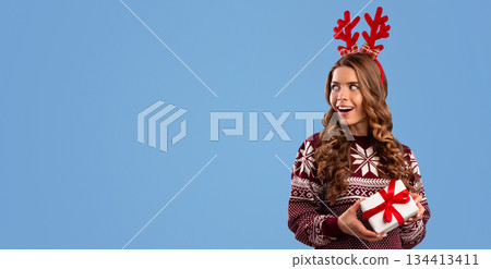 Sexy girl in warm sweater holding Christmas gift and looking at empty space, blue background. Panorama 134413411
