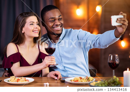 Happy Interracial Couple Taking Selfie On Smartphone During Romantic Date In Restaurant. Cheerful Black Man And White Woman In Love Smiling At Phone Camera, Celebrating Valentine's Day Together 134413416