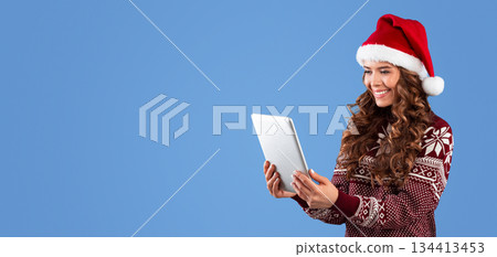 Christmas sale. Pretty lady in woolen sweater and Santa hat shopping online on tablet computer, blue background. Panorama 134413453