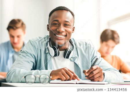 Modern Education Concept. Portrait of smiling African American male student sitting at desk in classroom at university, taking test or writing notes in his notebook, looking posing at camera 134413456