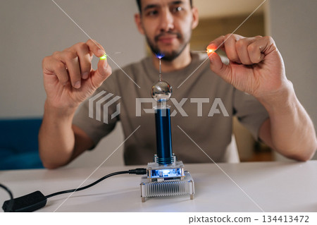 Bearded student male demonstrating mini tesla coil generating wireless power, excitingly lighting up small light bulbs through electromagnetic induction, experimenting with electricity and physics. Bearded student male demonstrating mini tesla coil generating wireless power, excitingly lighting up small light bulbs through electromagnetic induction, experimenting with electricity and physics. 134413472