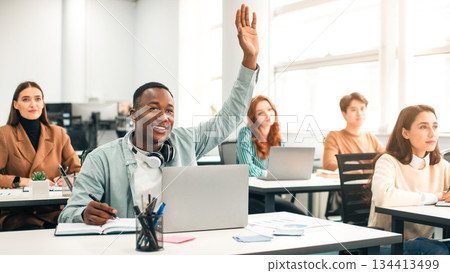 Education Concept. Smiling African American Male Student Raises Hand Up and Asks Lecturer a Question, Using Laptop Listening to Lecture at the University. Multi Ethnic Group of Modern Bright Students Education Concept. Smiling African American Male Student Raises Hand Up and Asks Lecturer a Question, Using Laptop Listening to Lecture at the University. Multi Ethnic Group of Modern Bright Students 134413499