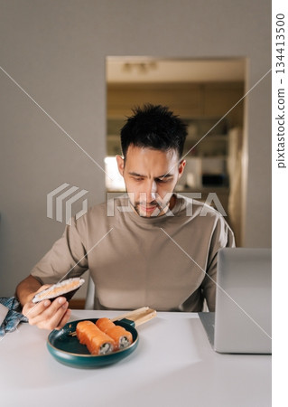 Vertical portrait of young man combining lunch with work from home, eating Japanese onigiri and sushi rolls while working on laptop computer sitting at desk, concept of remote employee lifestyle. 134413500