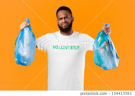 Discontented Black Volunteer Man Holding Two Plastic Garbage Bags Full Of Litter And Bottles Standing Over Yellow Studio Background. Waste Sorting, Junk Ecology Problem, Volunteering Work Concept 134413561